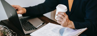 Person drinking coffee, looking at documents, and working on a laptop