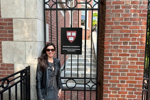Angela, a white woman with brown hair and sunglasses, poses in front of the gates of 51 Brattle Street, the Harvard Extension School building