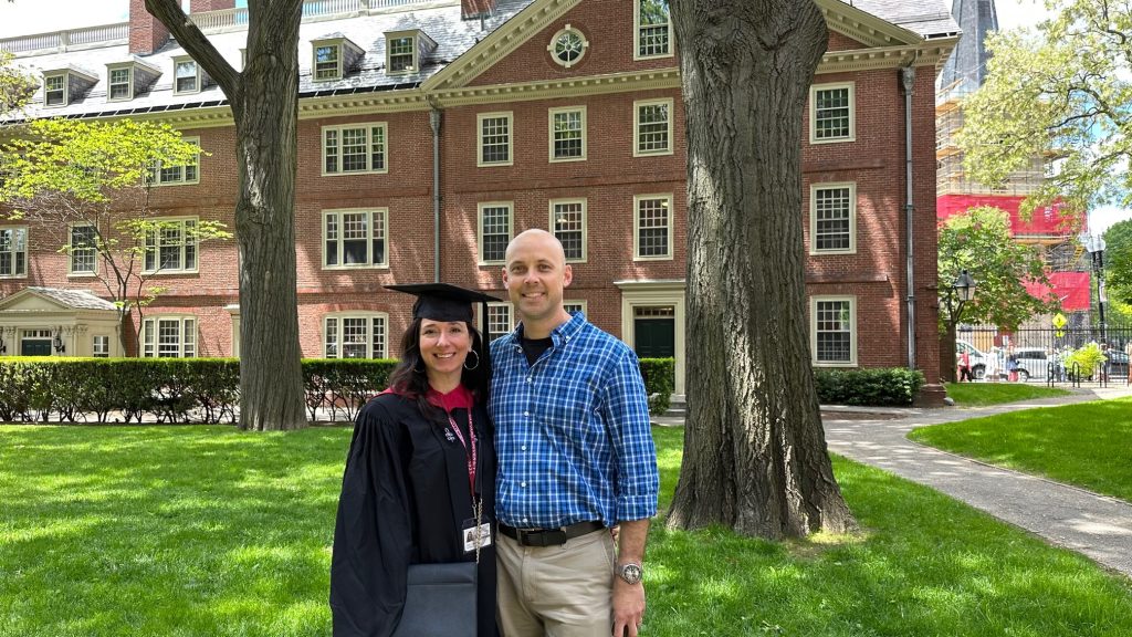 Angela, dressed in graduation cap and gown, and her husband pose smiling in front of a Harvard building