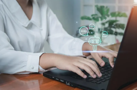 A person wearing a white shirt types on a laptop; the image of a robot with text bubbles is above their hands