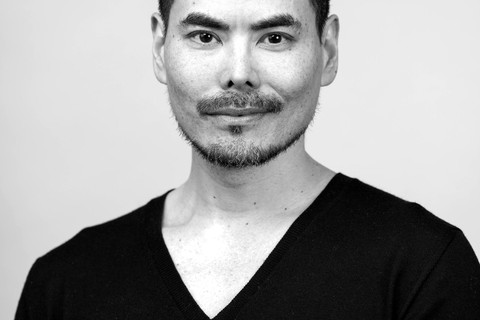 Black and white portrait of a person with short hair and a trimmed beard, wearing a black V-neck sweater, standing against a plain background and looking at the camera.