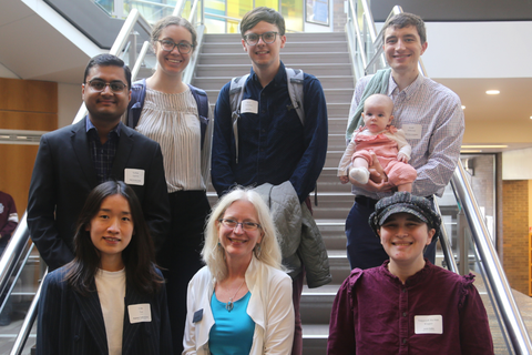 Group photo of seven adults, one holding an infant, gathered on a staircase inside a modern building.